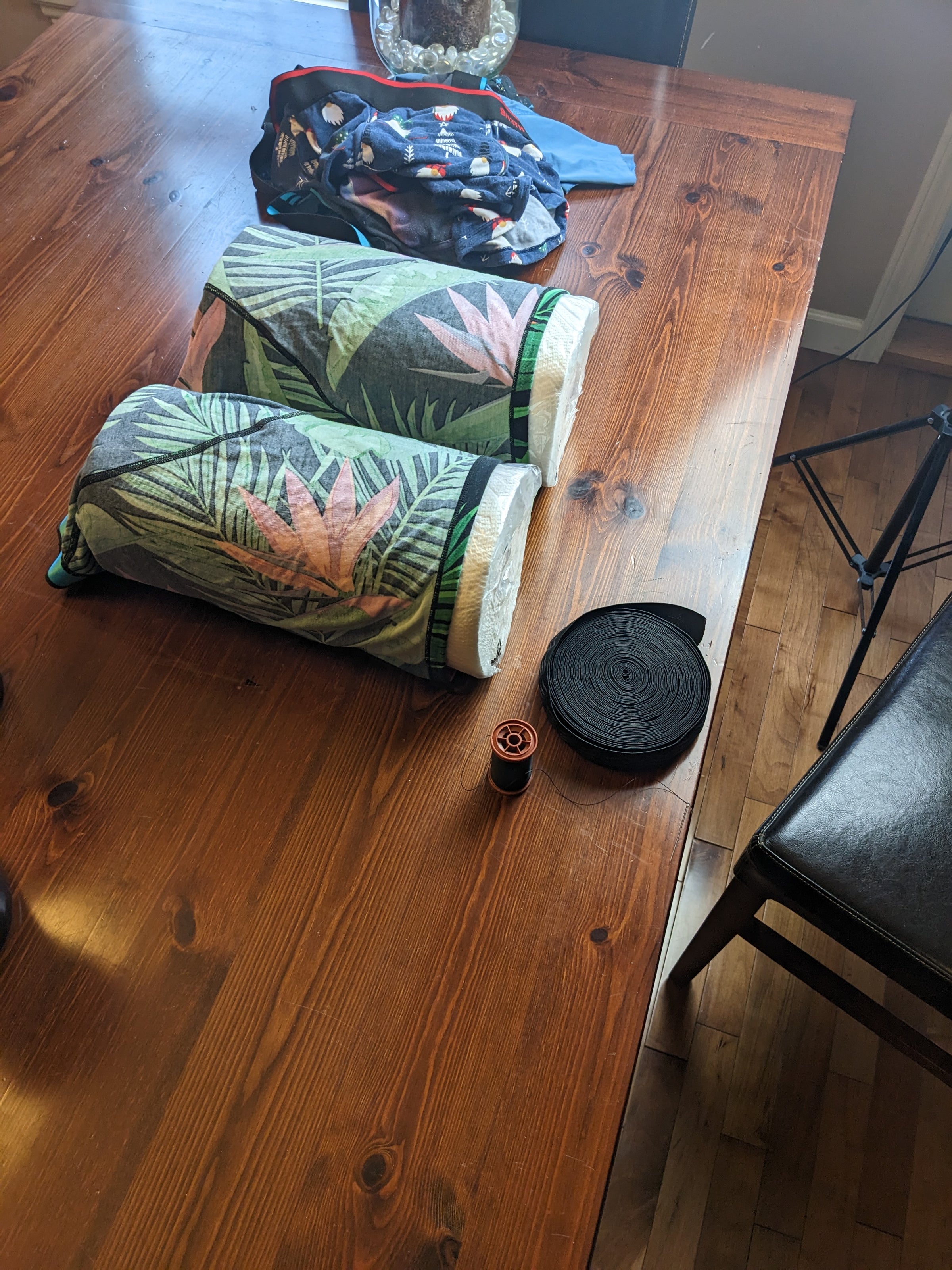 Hand-sewing experiments on a wooden dining table: printed fabric stitched around paper towel rolls, a coil of black band and a spool of thread