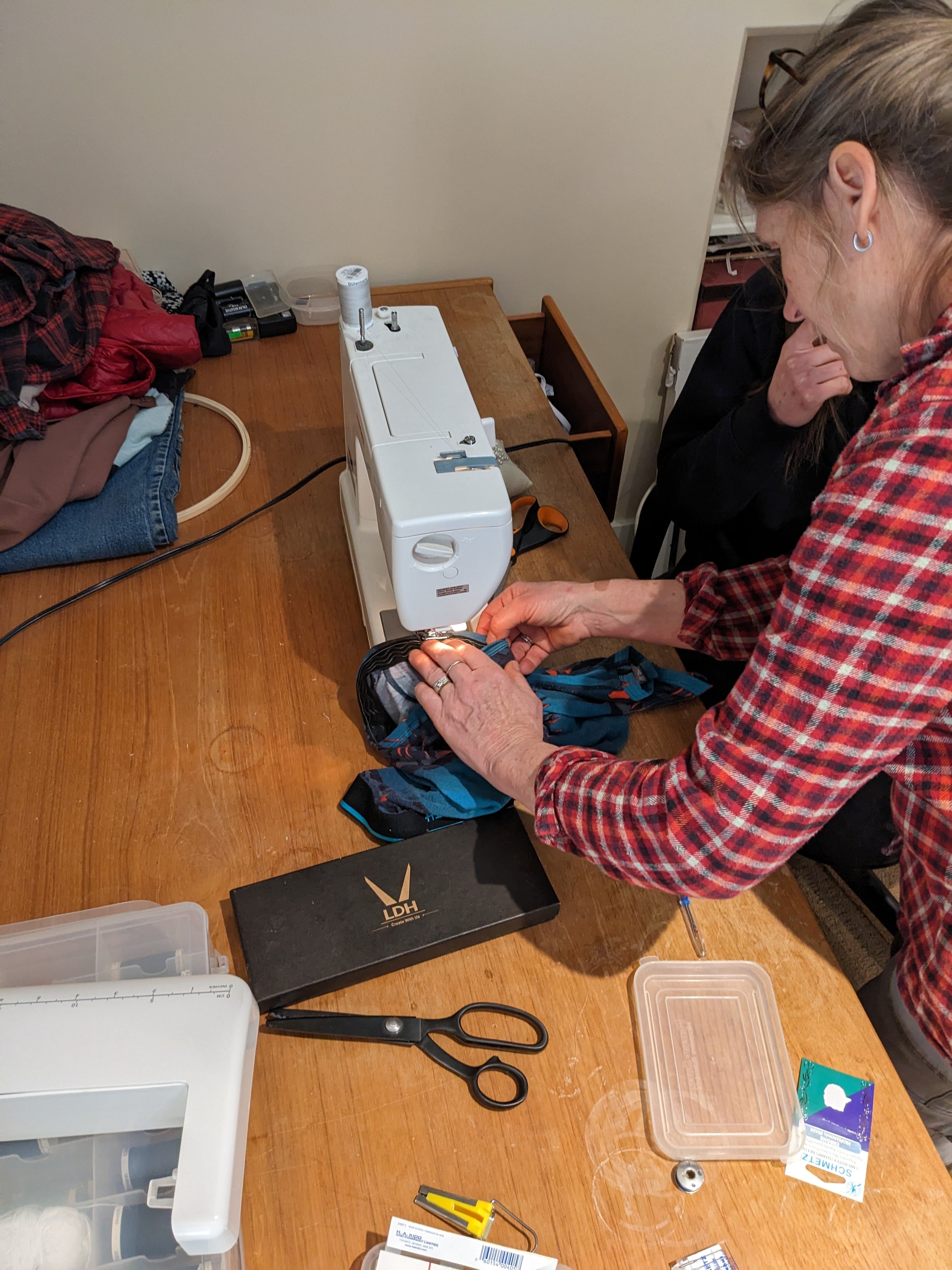 Hands guiding dark prototype fabric under the needle of a home sewing machine on a dining table, scissors and thread beside it