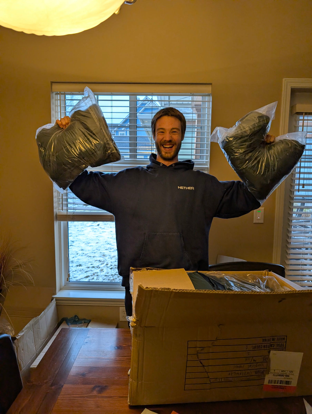 Stephen holding up two sealed bags of finished Nether boxer briefs over an open shipping carton, snow outside the window