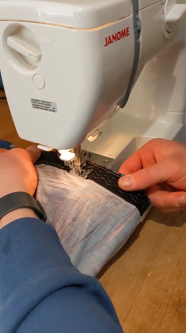 Hands feeding fabric under the needle of a Janome sewing machine on a wooden table