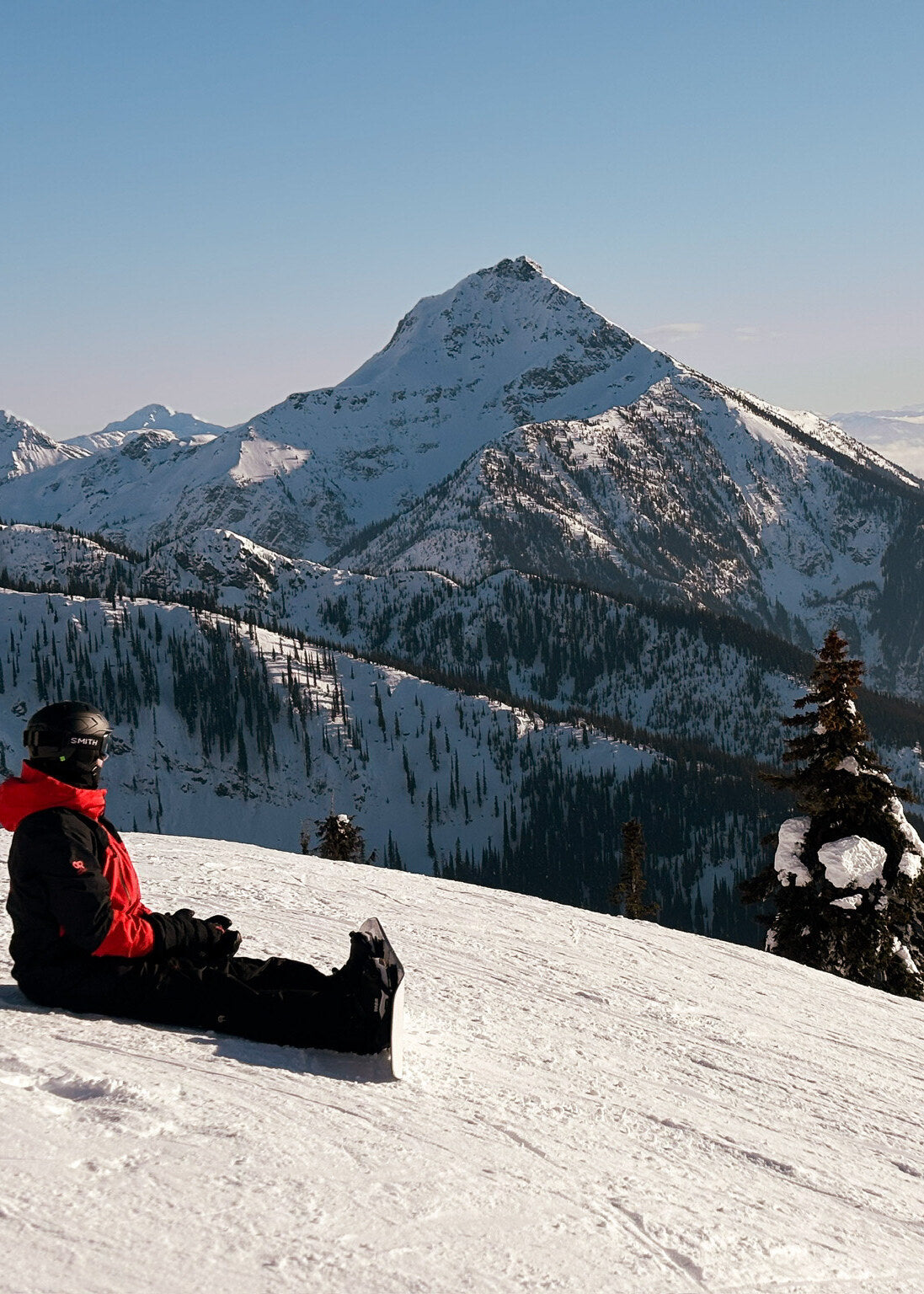 A snowboarder in a red jacket sitting on a groomed ridge at the top of a run, a snow-covered peak and a valley of cloud behind