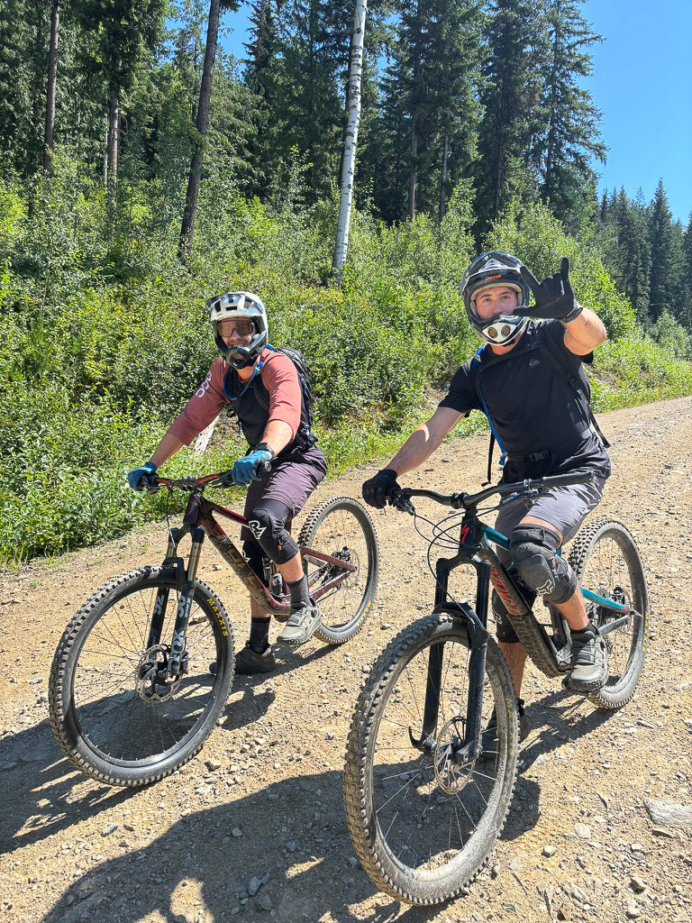 Two mountain bikers riding a gravel trail toward the camera through green forest under a blue sky